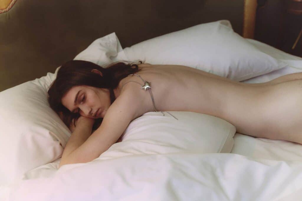A person with long hair lies face down on a bed with white pillows and sheets, appearing relaxed and partially covered, with a chain and pendant draped across their back.