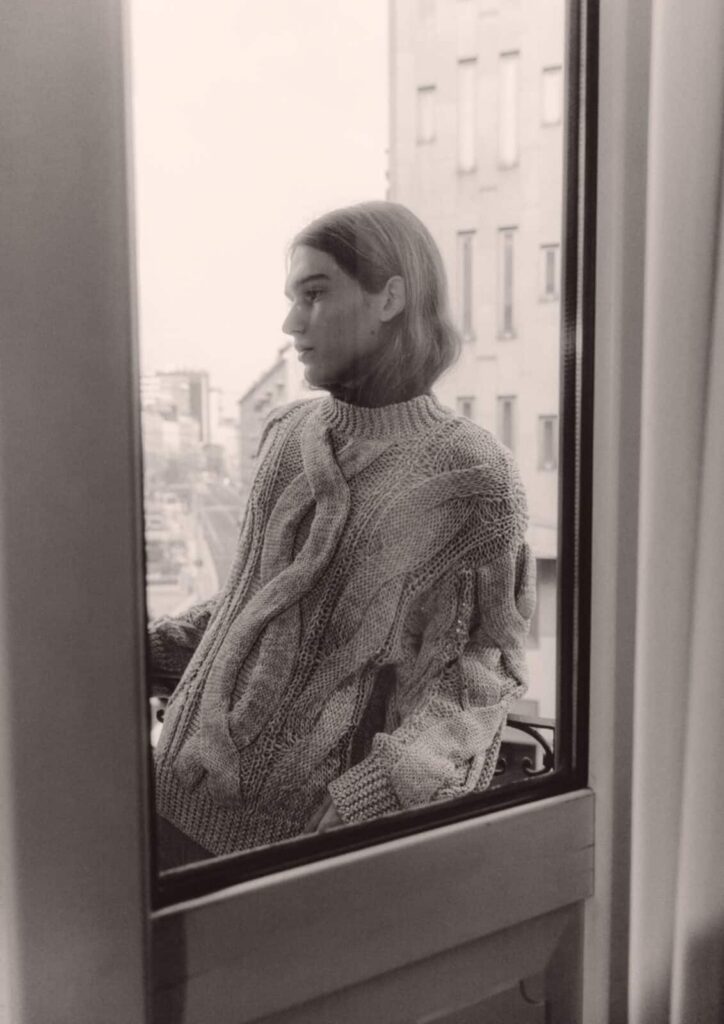 A person with long hair stands outside on a balcony, wearing a thick, cable-knit sweater, looking thoughtfully into the distance. The scene is viewed through a window, and the cityscape is visible in the background.
