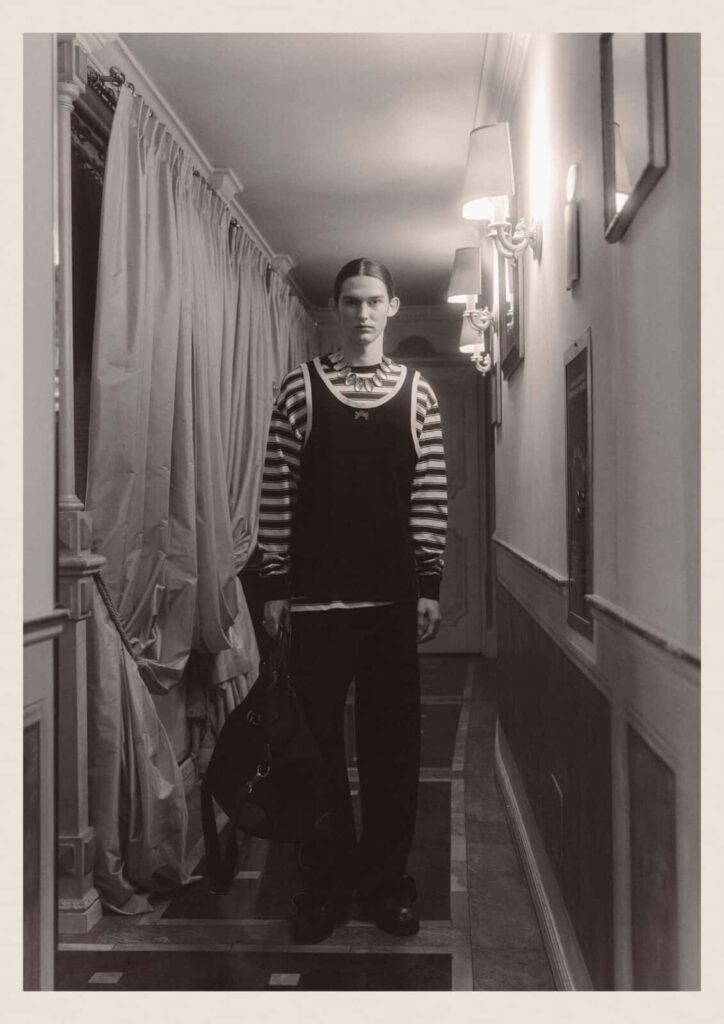 A person with long hair stands in a narrow, dimly lit hallway, wearing a striped long-sleeve shirt layered under a dark tank top and loose trousers, holding a large bag in one hand. The scene has a vintage, monochrome look.