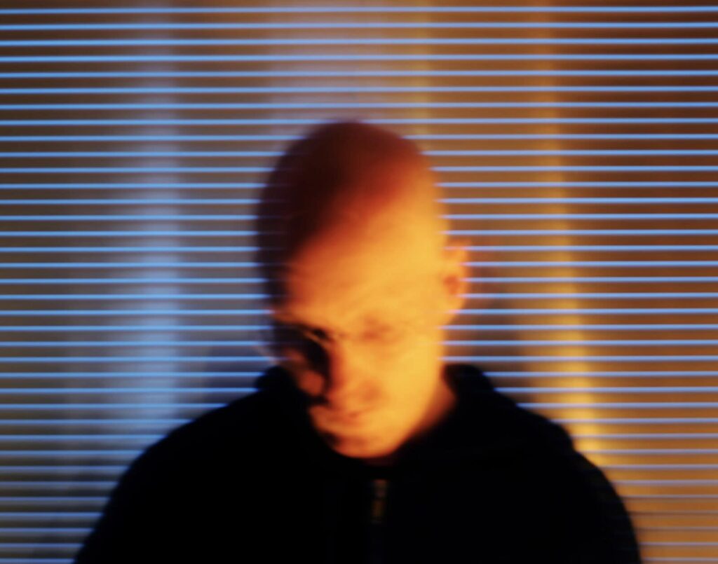 A blurry image of Ben Kreukniet person with glasses looking down, set against a background of horizontal window blinds with blue and orange lighting.