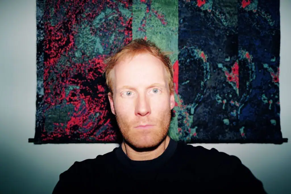 A man with light skin and reddish hair stares directly at the camera, illuminated by a flash. Behind him is a colorful, abstract tapestry with green, red, and dark blue patterns.