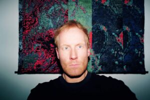 A man with light skin and reddish hair stares directly at the camera, illuminated by a flash. Behind him is a colorful, abstract tapestry with green, red, and dark blue patterns.
