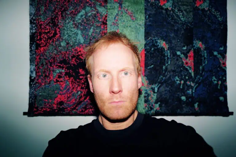 A man with light skin and reddish hair stares directly at the camera, illuminated by a flash. Behind him is a colorful, abstract tapestry with green, red, and dark blue patterns.