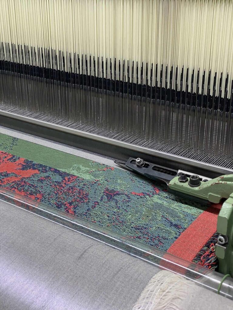 A close-up view of an industrial weaving loom creating a colorful patterned textile with red, green, and blue threads. Numerous vertical threads are aligned in the background.