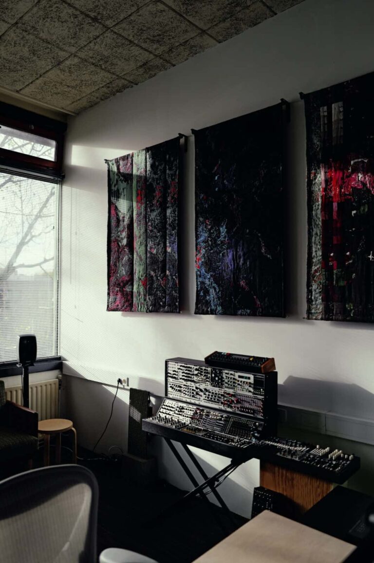 A music studio corner with a large modular synthesizer on a stand beneath three abstract wall hangings. Sunlight streams through a window, illuminating part of the room. A stool and chair are also visible.