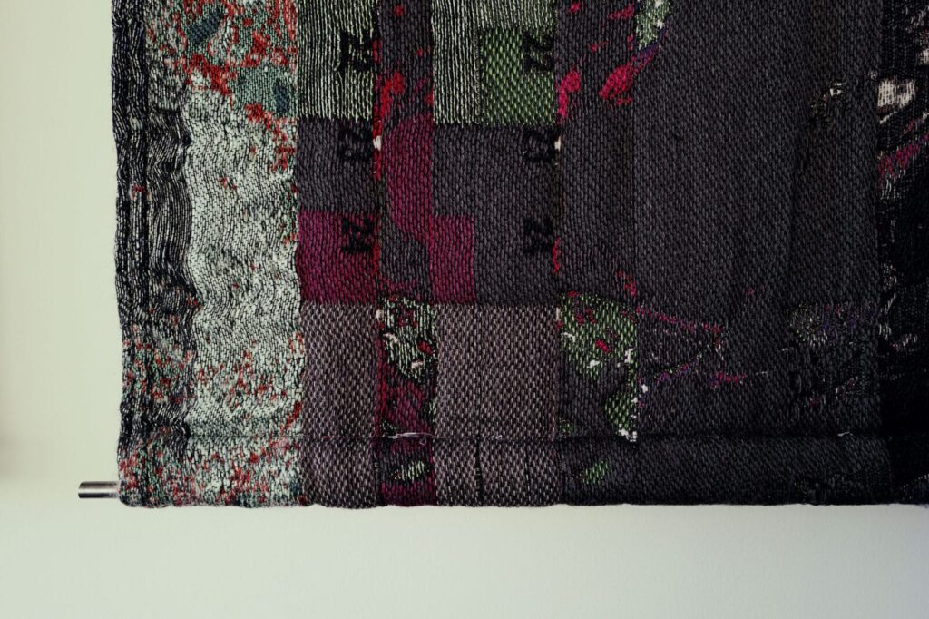 A close-up of a textured, woven textile hanging on a wall, featuring patchwork squares in dark shades of gray, green, red, and black, with some metallic threads and intricate patterns, artwork by Ben Kreukniet.