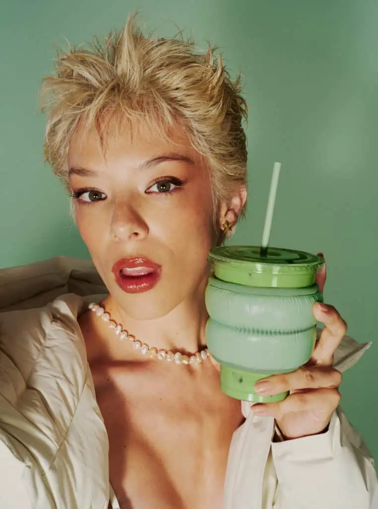 A person with short, tousled blonde hair poses against a green background, holding a green Blank Street cup with a straw. They wear a white jacket off the shoulder, a pearl necklace, and have glossy lips.
