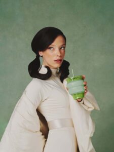 A woman with dark, styled hair and a serious expression wears a light, futuristic outfit and holds a green Blank Street collapsible cup with a straw against a plain green background.
