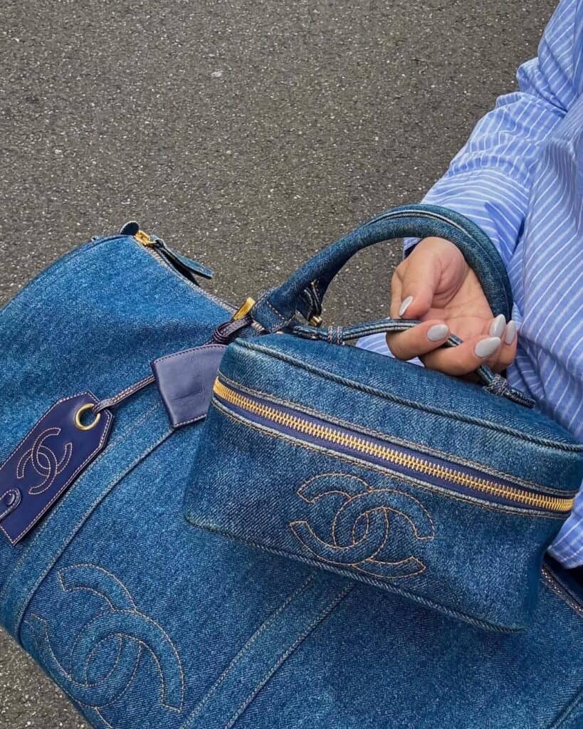 A person wearing a blue striped shirt holds two matching blue denim Chanel bags with gold zippers and logos, one large duffle and one small case, against a gray pavement background.