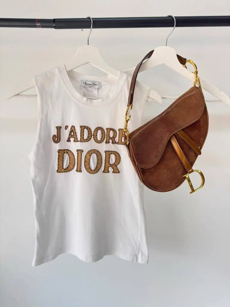 A white HIVE sleeveless top with JADORE DIOR in gold lettering hangs on a hanger next to a brown leather Dior saddle bag with gold hardware, both displayed on a black clothing rack.