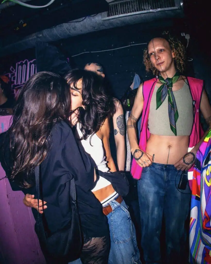Three people stand in a dimly lit, colorful nightclub. Two are kissing intimately in the foreground, while a third person in a bright vest, scarf, and crop top stands nearby, looking toward the camera.