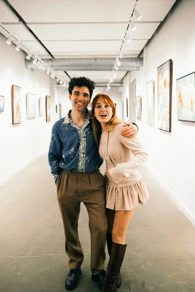 Arden Rose & Adrian Bliss stand arm in arm in an art gallery hallway, surrounded by paintings on white walls. The man wears brown pants and a blue shirt, while the woman wears a light pink dress and brown boots.