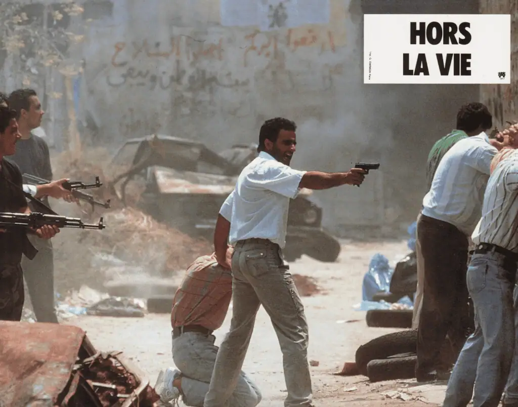 A tense street scene shows armed men aiming guns; one man in the center points a pistol, while others hold rifles. Two people are kneeling with hands behind their heads. A poster reads Hors la vie E in bold letters. The background is chaotic.