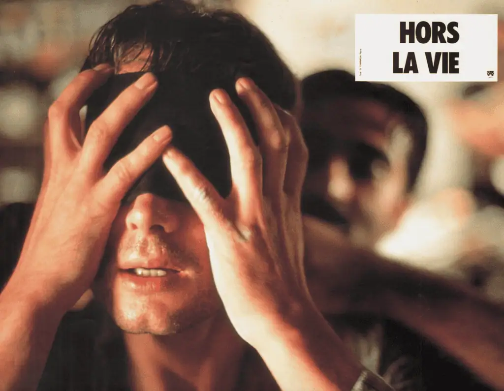 A close-up of a man with his hands covering his own eyes, looking tense. Another person is slightly blurred in the background. Text in the top right corner reads: Hors la vie.