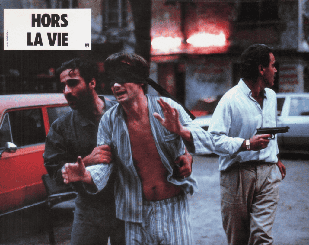 A blindfolded man in pajamas is escorted by two men; one holds his arm and the other points a handgun, with a red car and blurred city lights in the background. Hors la vie is written in bold text at the top left.