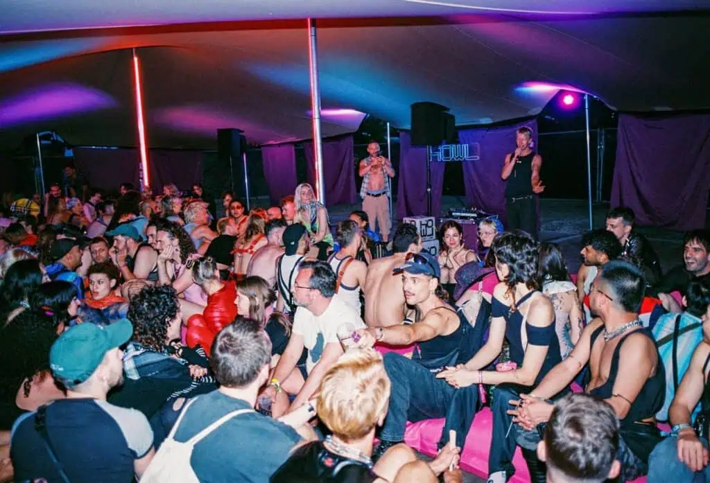 A large group of people in revealing and creative outfits sits closely together under a tent with colorful Valentine lighting. Some people look toward a stage where two individuals are speaking or hosting the event.