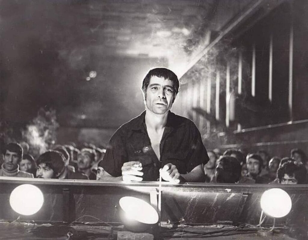 A man stands at a table with spotlights, looking forward in a dimly lit room filled with seated people in the background. The photo is black and white, creating a dramatic and intense atmosphere.
