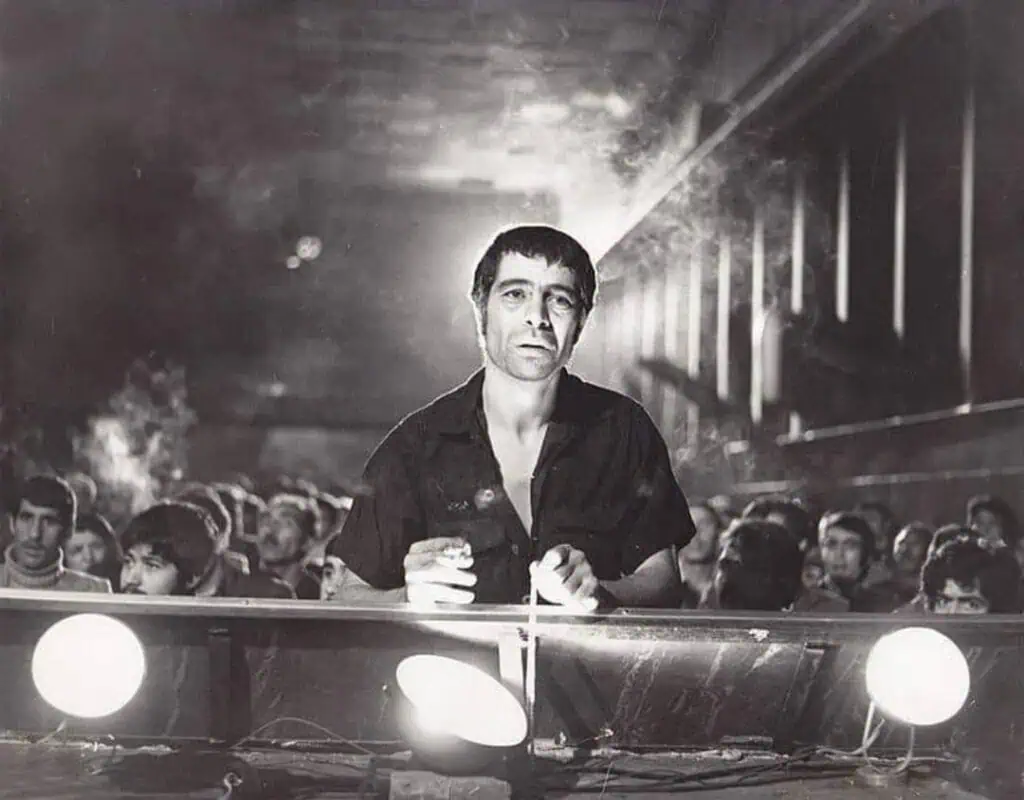 Frames of Memory at the Barbican: A man stands at a table with spotlights, looking forward in a dimly lit room filled with seated people in the background. The photo is black and white, creating a dramatic and intense atmosphere.