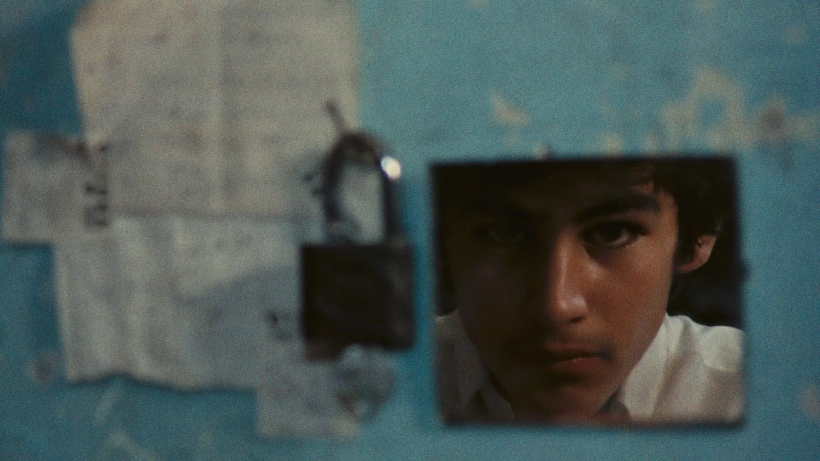 A young person’s face is partially reflected in a small, square mirror on a blue wall with faded papers and a black padlock hanging nearby. The expression appears serious and contemplative.