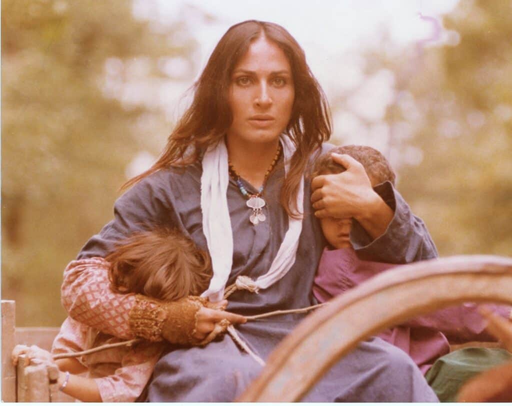 A woman in a blue dress and white scarf sits on a wooden cart, protectively holding two children close to her. One child leans on her arm while the other is partially hidden in her embrace. The scene is outdoors and has a soft focus.