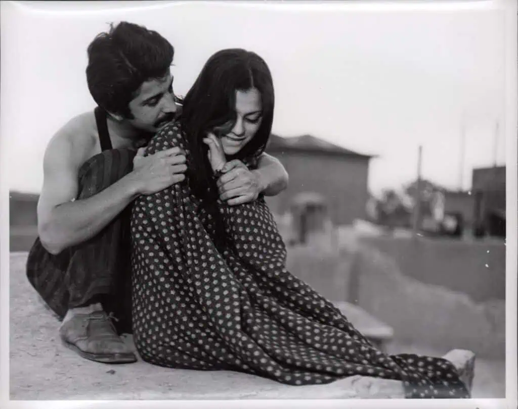 A young man sits behind a young woman, gently holding her shoulders. The woman looks down with a soft smile, draped in a patterned fabric. They are outdoors, with blurred buildings in the background. The photo is black and white.