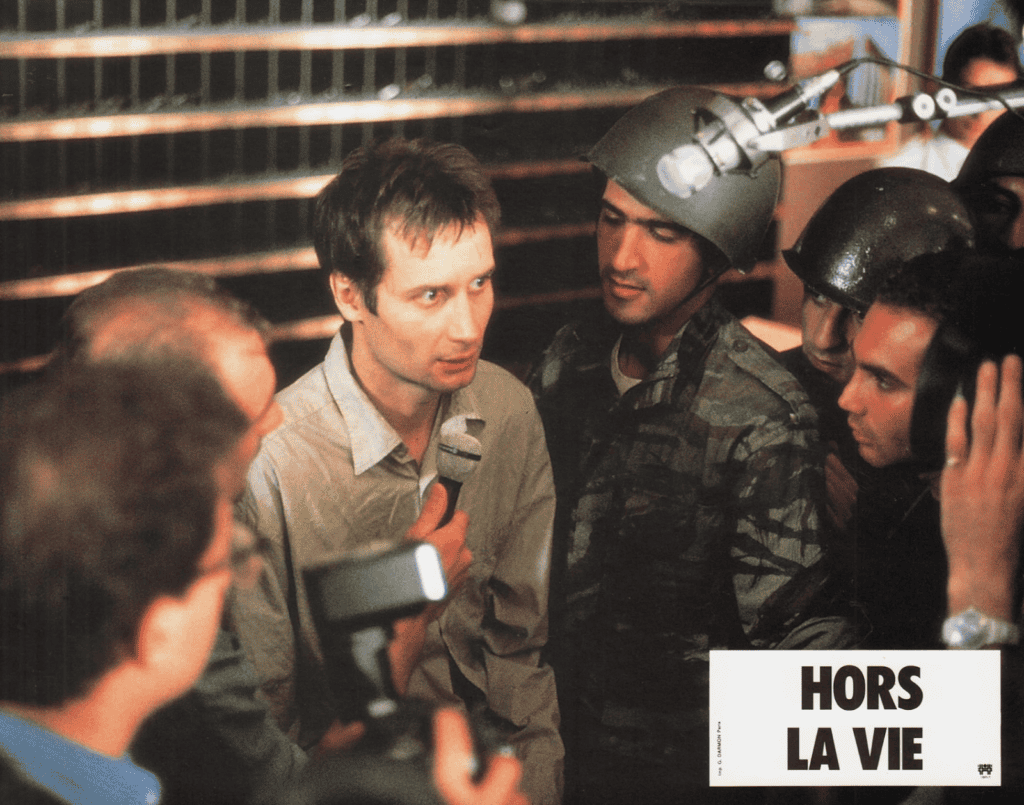 A man holding a microphone is surrounded by soldiers in helmets and journalists with cameras and recording equipment. The text HORS LA VIE appears in the bottom right corner.
