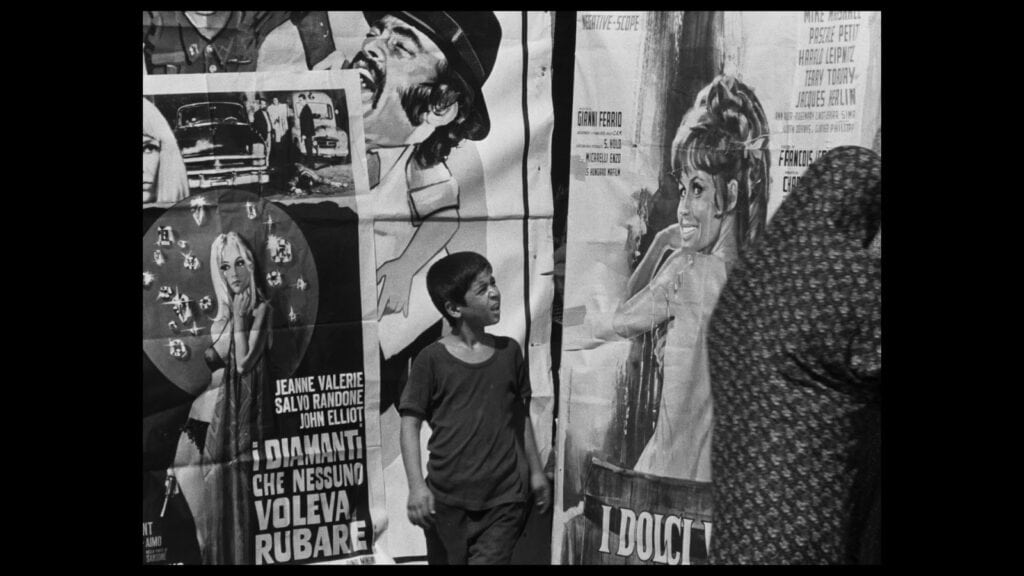 A young boy looks up at colorful movie posters on a city wall; one features a smiling woman, another a man in a hat. An adult, partially seen, walks past in the foreground. The scene is in black and white.