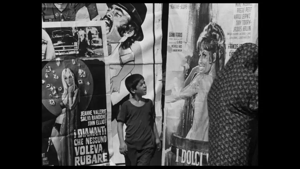Frames of Memory at the Barbican: A young boy looks up at colorful movie posters on a city wall; one features a smiling woman, another a man in a hat. An adult, partially seen, walks past in the foreground. The scene is in black and white.