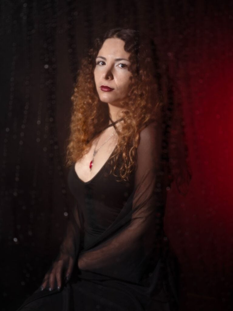 A woman with long curly hair wearing a black dress and a red pendant necklace sits against a dark background with a sheer black curtain and red lighting on one side.