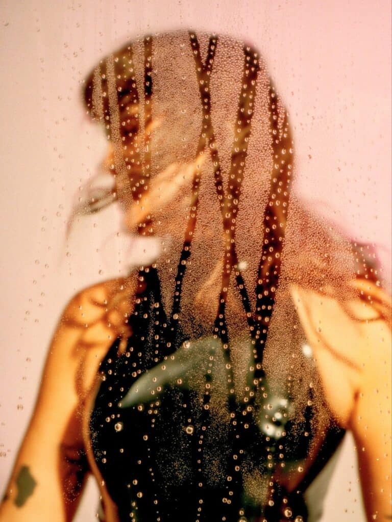 A person in a sleeveless black top stands behind a foggy, wet glass pane with water droplets, creating a blurred effect. Their face and features are indistinct, and light filters through the scene.