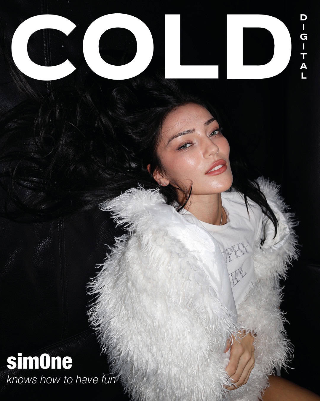 A woman with long dark hair, wearing a white furry coat and white top, lies on a black surface and looks at the camera. The magazine cover reads COLD DIGITAL and simOne knows how to have fun.