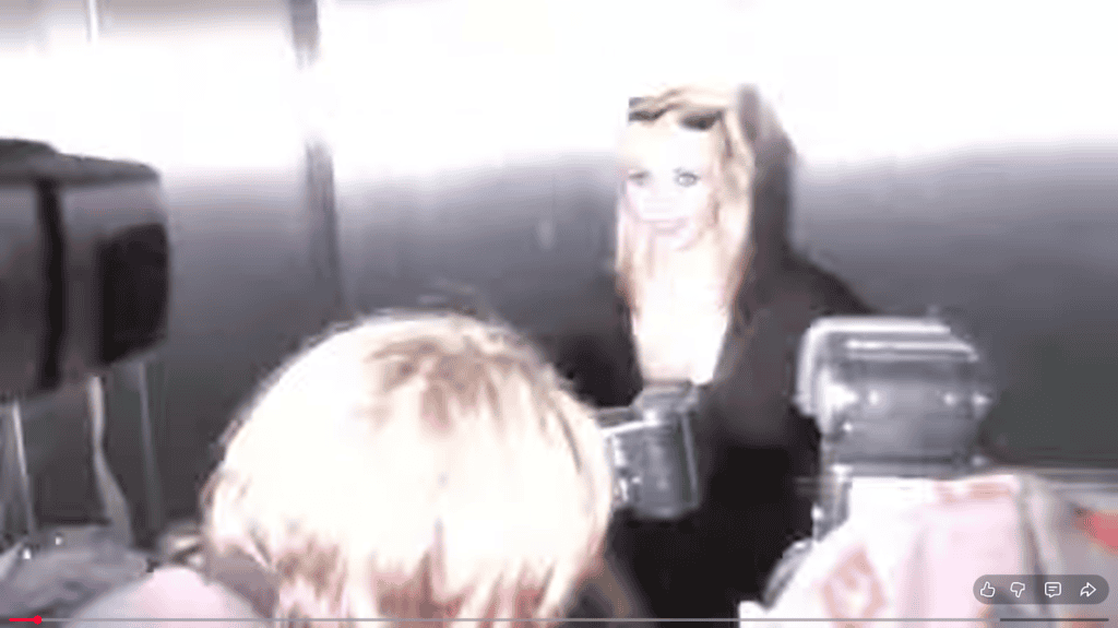 A woman with blonde hair and sunglasses on her head stands in front of a bright camera flash, surrounded by photographers with visible cameras and flashes.