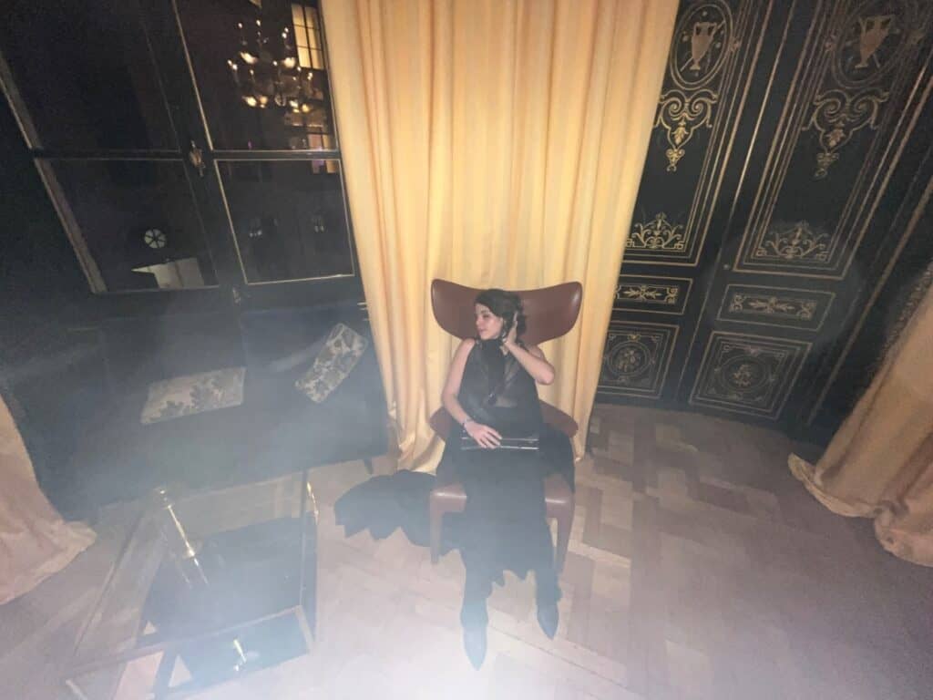 A woman in a black dress sits on a brown chair in a dimly lit, ornate room with tall yellow curtains, black and gold decorated walls, and wooden parquet flooring. She is looking to the side and holding a small purse.