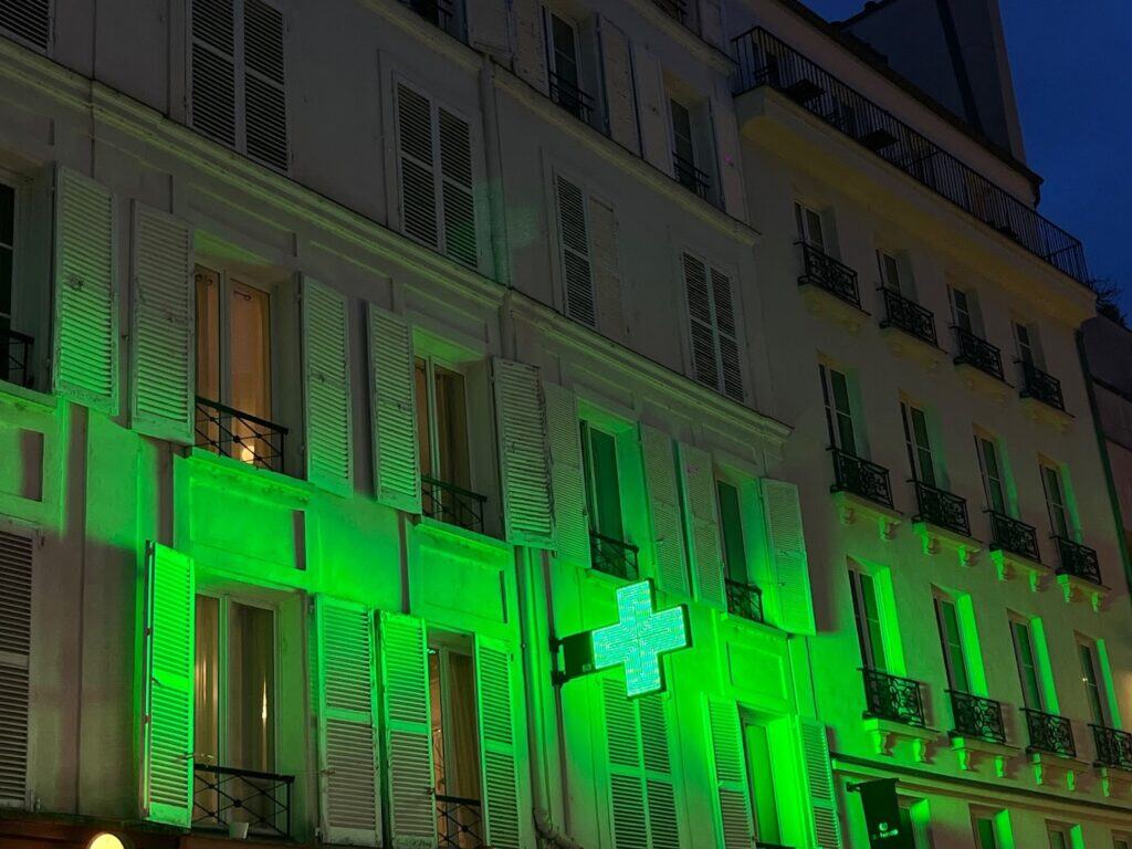 A building with white shutters and several balconies is illuminated by green lights at dusk. A glowing green cross sign, indicating a pharmacy, is mounted on the facade.