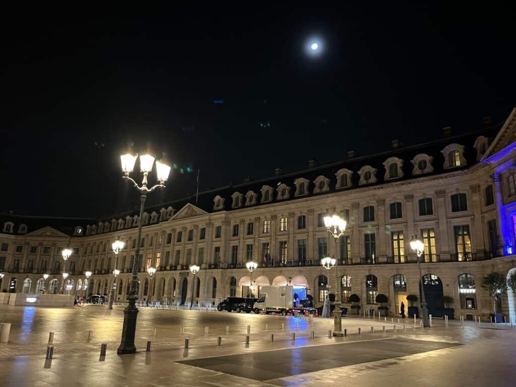 A grand, historic building is illuminated at night in a large, empty plaza. Street lamps glow warmly and the full moon shines in the dark sky above, adding to the tranquil atmosphere.