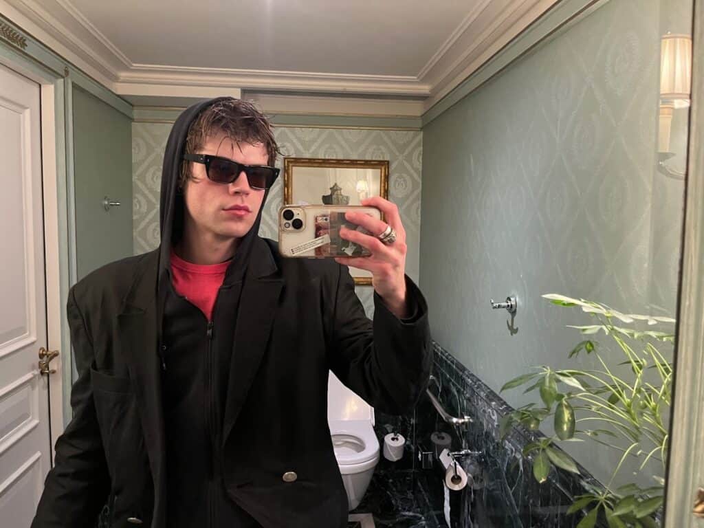 A young person wearing sunglasses, a black jacket, and a hood takes a selfie in a well-lit bathroom with green walls, a plant, and a toilet visible in the background.
