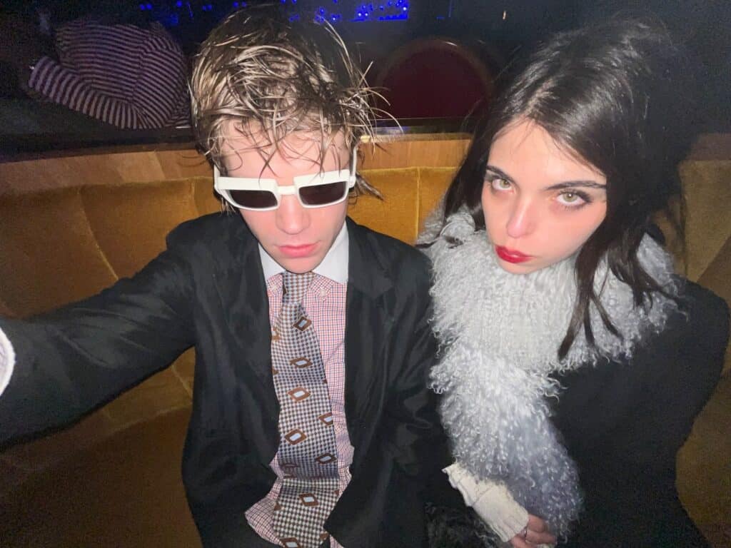 Two young adults sit closely together on a velvet bench in a dimly lit setting. One wears white sunglasses and a suit with messy hair; the other has long dark hair, a fluffy scarf, and red lipstick. Both have serious expressions.