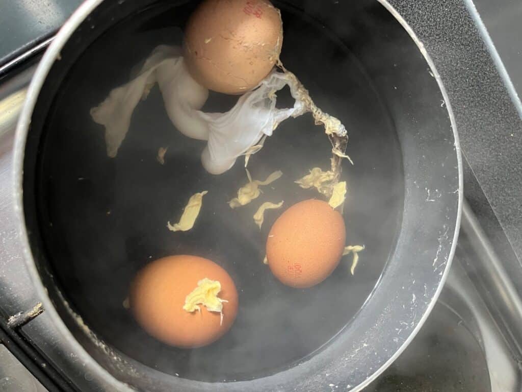 Three brown eggs are boiling in a pot of water, with some egg whites leaking out from one cracked egg. Pieces of cooked egg white float in the water. Steam rises from the pot.