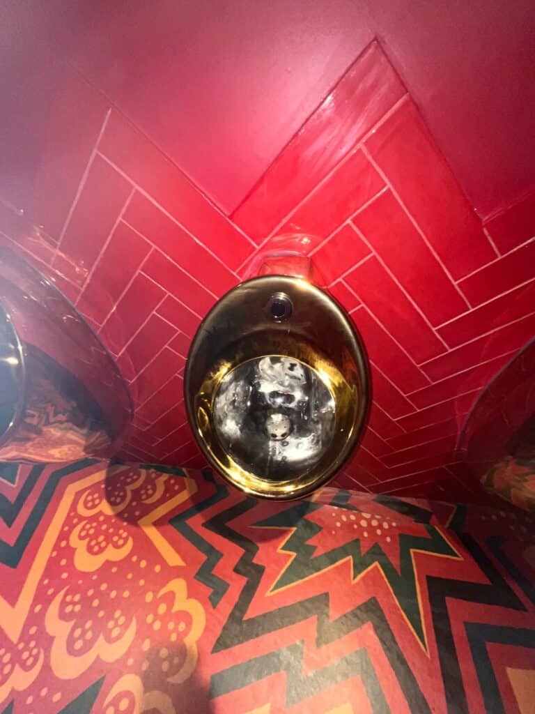 A gold urinal mounted on a red tiled wall with zigzag patterns, above a colorful, patterned floor featuring starbursts and floral designs in red, black, and orange tones.