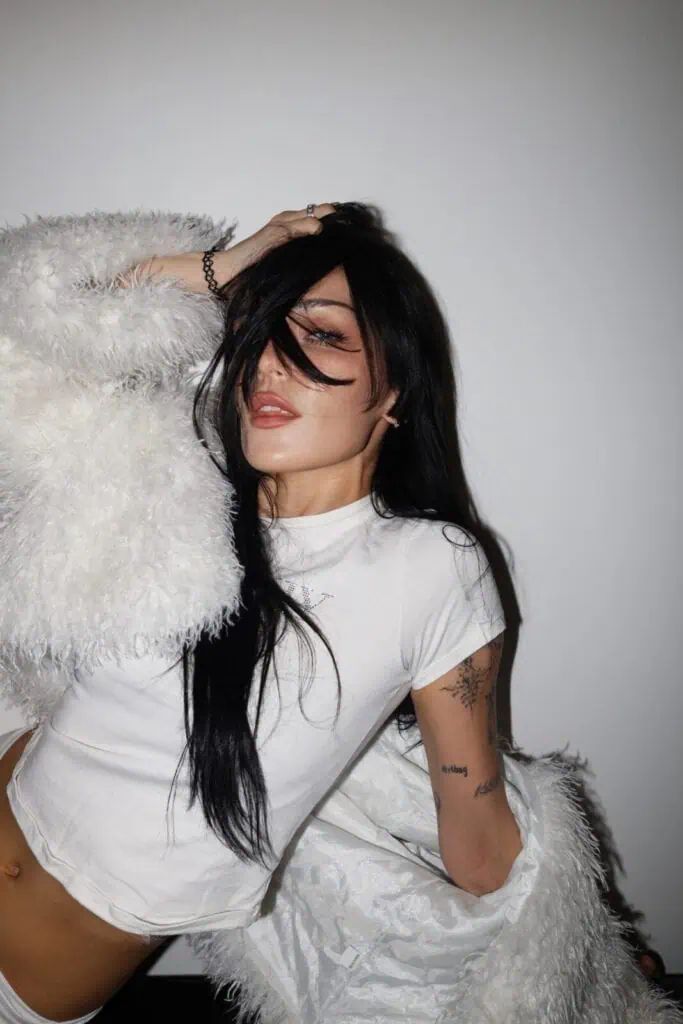 A woman with long black hair poses against a plain white wall, wearing a white furry jacket and crop top. Channeling her signature style from the Sim0ne interview, her hair partially hides her face as she reveals tattoos on her arm.
