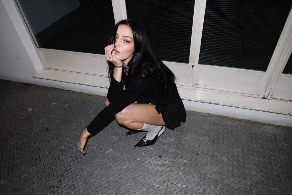 A woman with long dark hair squats on a metal floor indoors, wearing a black outfit, white socks, and black shoes. Channeling the poise of a sim0ne interview, she touches her face and gazes confidently towards the camera.