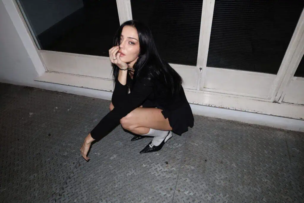 Simone Murphy with long dark hair squats on a metal floor indoors, wearing a black outfit, white socks, and black shoes. Channeling the poise of a sim0ne interview, she touches her face and gazes confidently towards the camera.