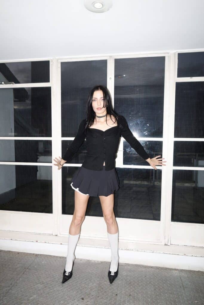 A woman with long dark hair stands indoors against a window, dressed as if ready for a sim0ne interview. In a black cardigan, short skirt, knee-high white socks, and heels, she faces forward, arms outstretched to touch the wall.