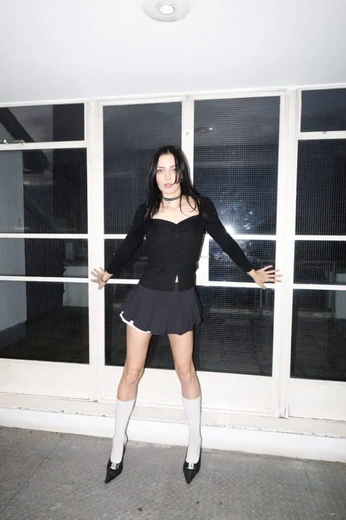 A woman with long dark hair stands indoors against a window, dressed as if ready for a sim0ne interview. In a black cardigan, short skirt, knee-high white socks, and heels, she faces forward, arms outstretched to touch the wall.