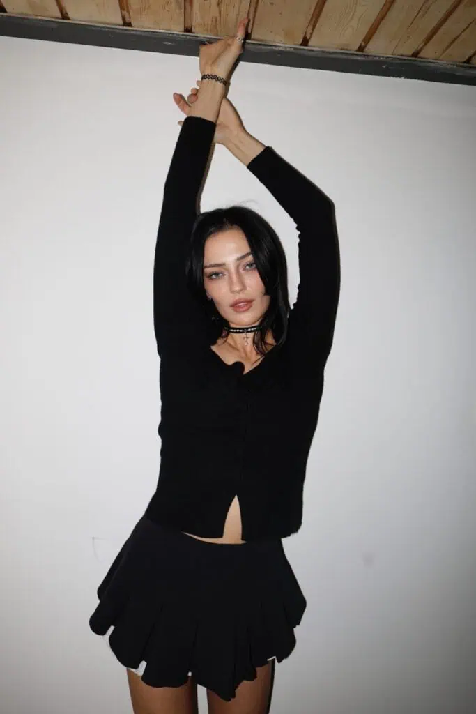 Simone Murphy with dark hair, channeling simone murphy, stands against a white wall in a black long-sleeve top and skirt, arms raised above her head, looking at the camera with a neutral expression.