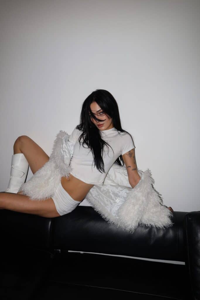 A woman with long black hair poses on a black couch, channeling Simone Murphy’s iconic style from the Sim0ne interview. She wears a white crop top, shorts, knee-high boots, and a fluffy coat, leaning back with one leg up against a plain white wall.