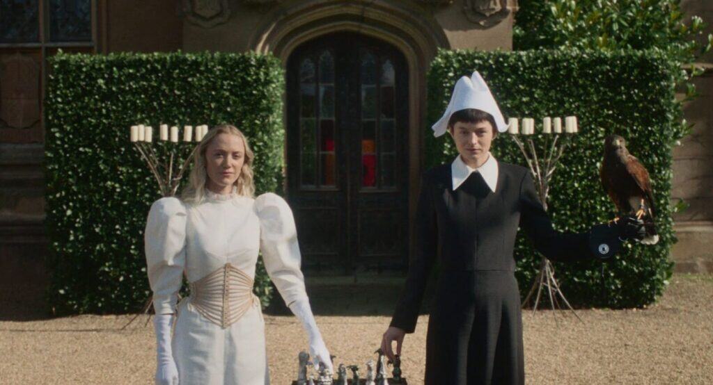 Two women stand side by side outdoors: one in a white dress with puffed sleeves and gloves, the other in a black dress and white bonnet, holding a bird of prey. A chessboard sits in front of them, with a mansion and greenery behind.