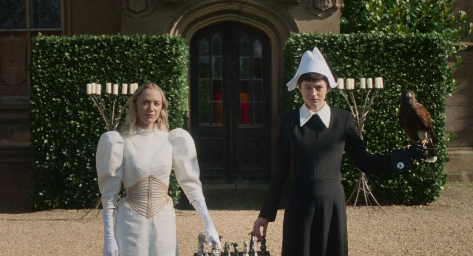 Two women stand side by side outdoors: one in a white dress with puffed sleeves and gloves, the other in a black dress and white bonnet, holding a bird of prey. A chessboard sits in front of them, with a mansion and greenery behind.