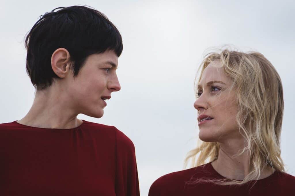 Two women wearing matching red tops stand side by side outdoors, gazing seriously at each other against a pale, cloudy sky. One has short dark hair, the other has long blond hair.