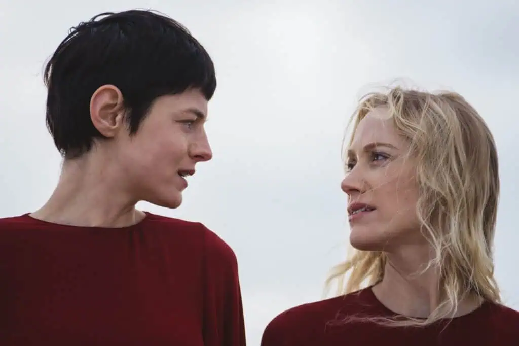 Two women wearing matching red tops stand side by side outdoors, gazing seriously at each other against a pale, cloudy sky. One has short dark hair, the other has long blond hair. Still from "It’s an Absurd World" directed by Julia Jackman.